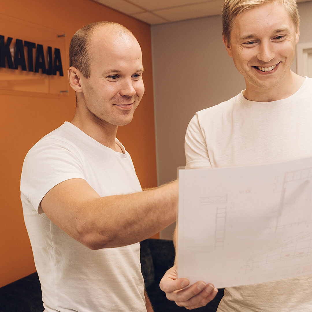 Kaksi miestä katsoo hymyillen rakennuspiirustuksia toimistossa oranssin seinän edessä